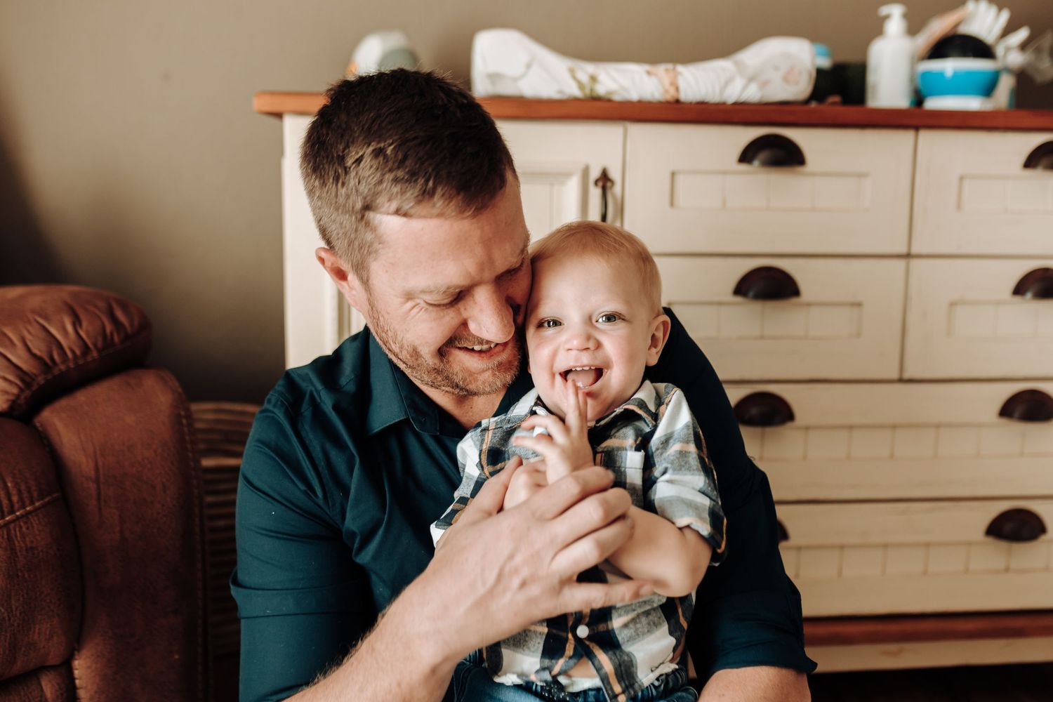 A parent and baby share a joyful moment together while sitting in front of a white dresser at home.