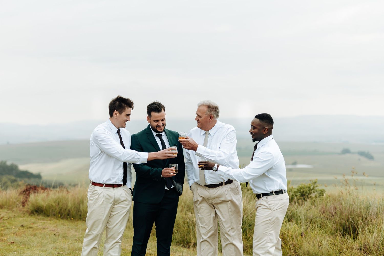 A group of people in formal attire celebrating together in a scenic outdoor field with rolling hills in the background.