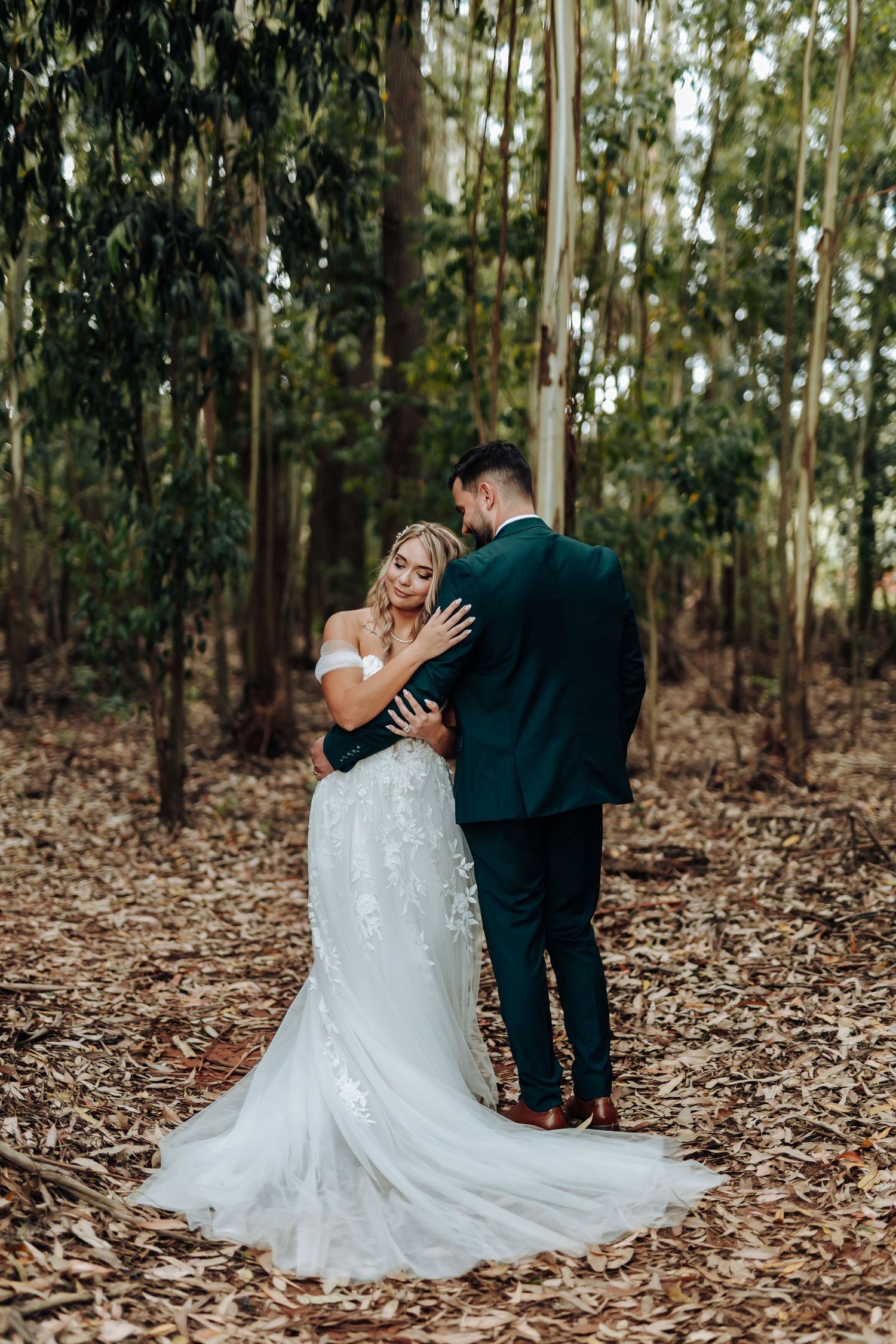 Newlyweds share an intimate moment among eucalyptus trees while the bride's lace gown spreads beautifully on forest floor.