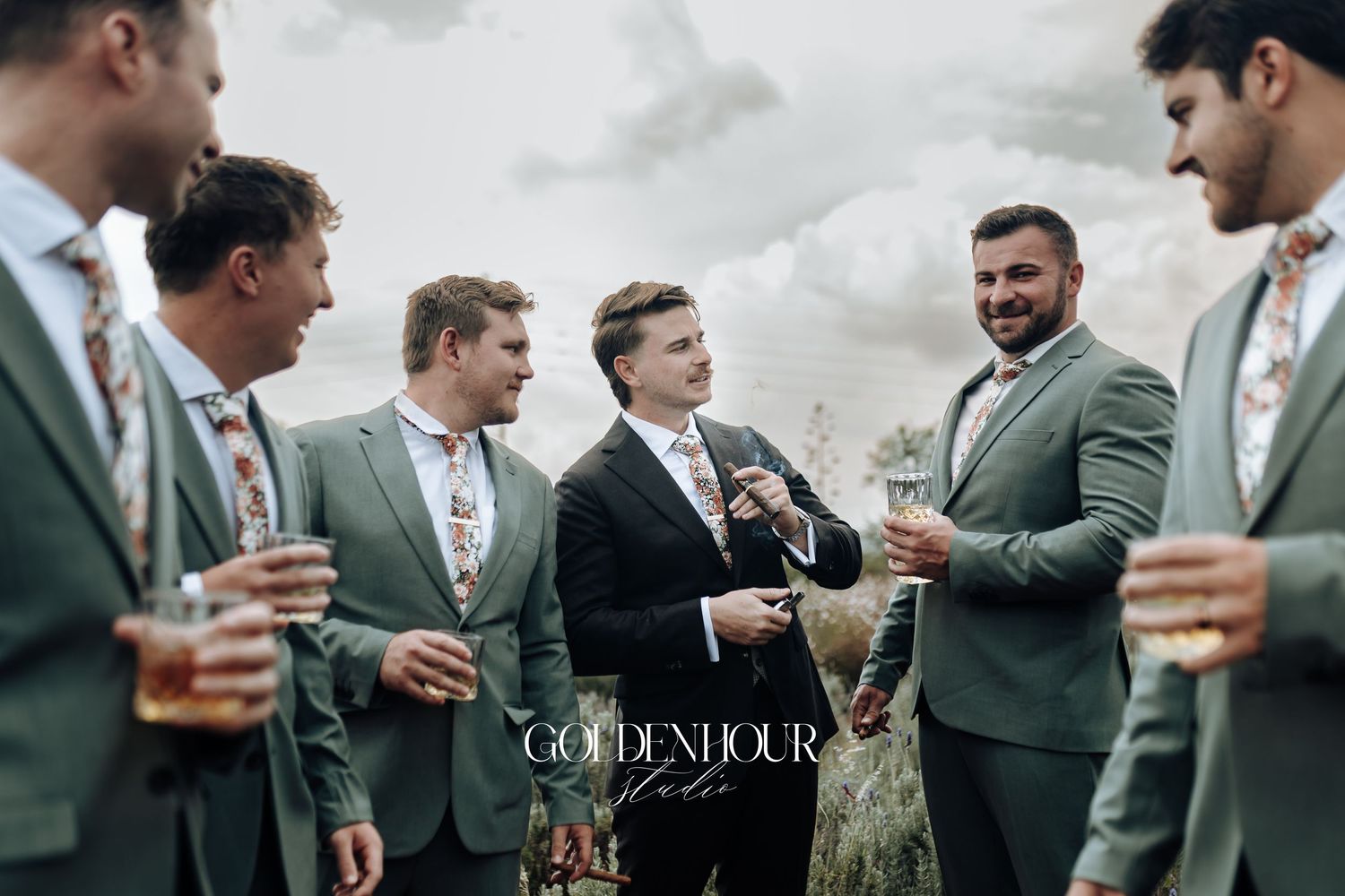 Groomsmen in olive green suits share drinks and laughs during an outdoor wedding celebration.