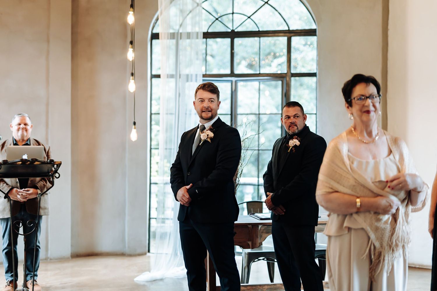Wedding party poses in elegant white venue with arched windows and hanging pendant lights.