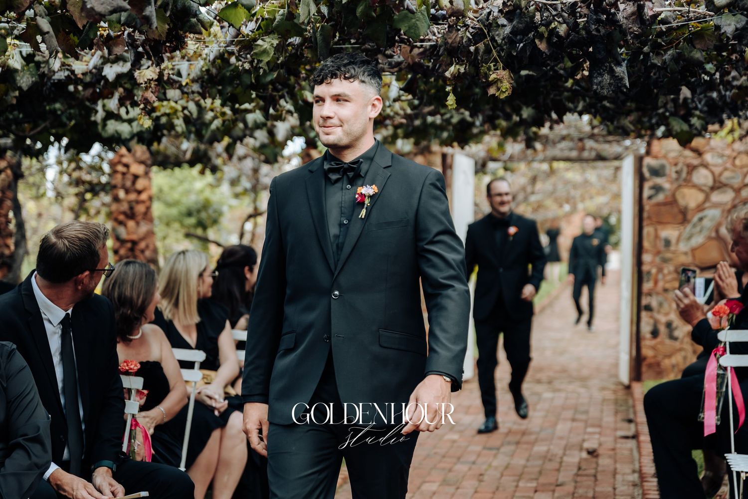 A guest in a dark suit stands among others at an outdoor garden wedding ceremony venue.