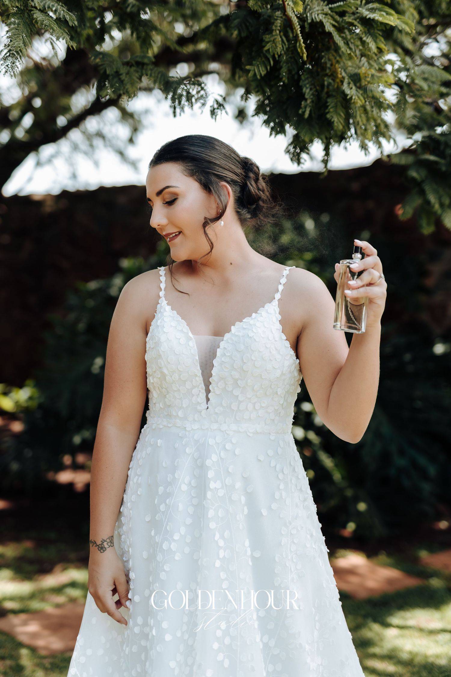 A person in a white beaded wedding dress holds perfume bottle while standing under evergreen branches.