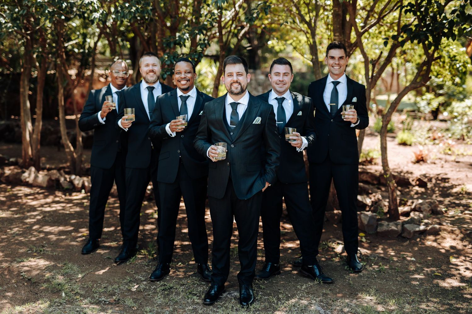 Groomsmen in black suits share laughter and drinks during outdoor wedding photo session in wooded setting.