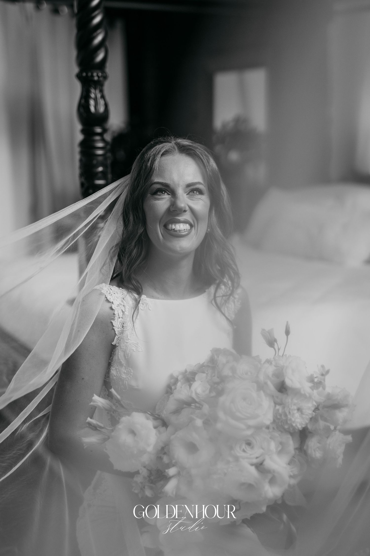 A joyful bride in a white dress holds a beautiful bouquet while wearing a flowing veil in a black and white portrait.