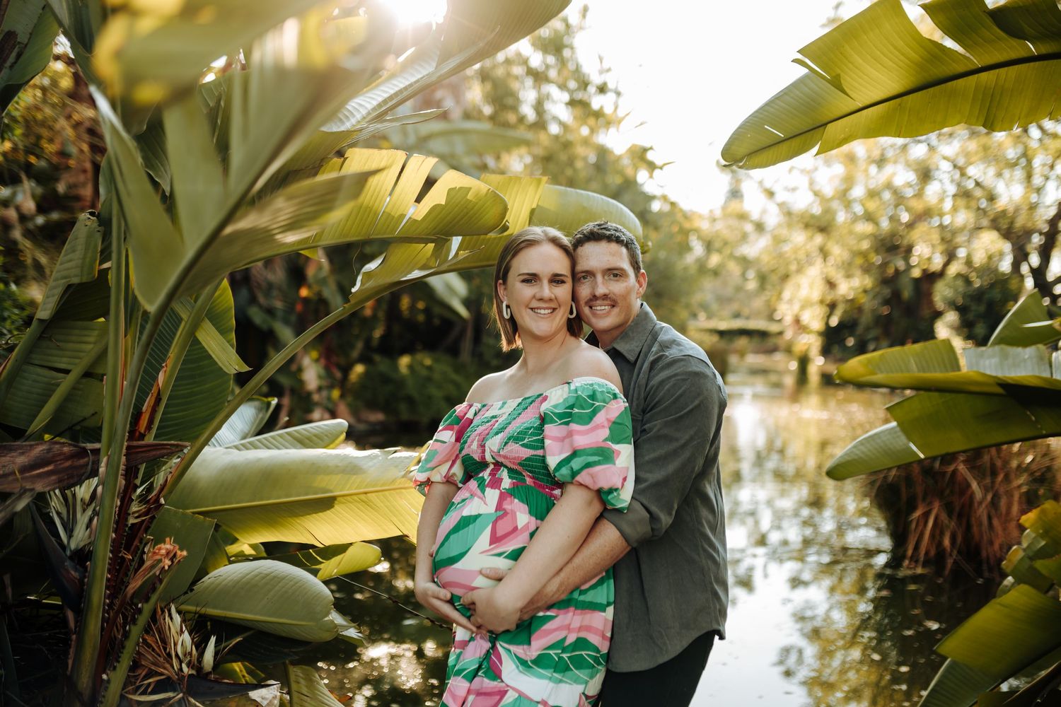 Romantic couple embraces lovingly under tropical foliage during sunset photoshoot at scenic outdoor location.