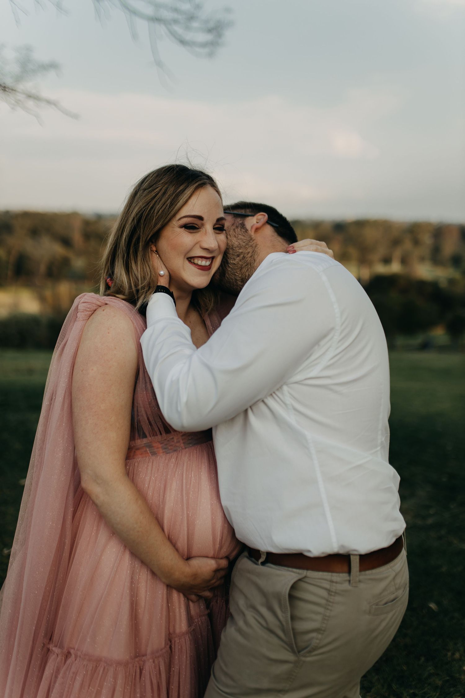 A couple in formalwear shares an intimate moment and laughter together in a scenic outdoor field at sunset.