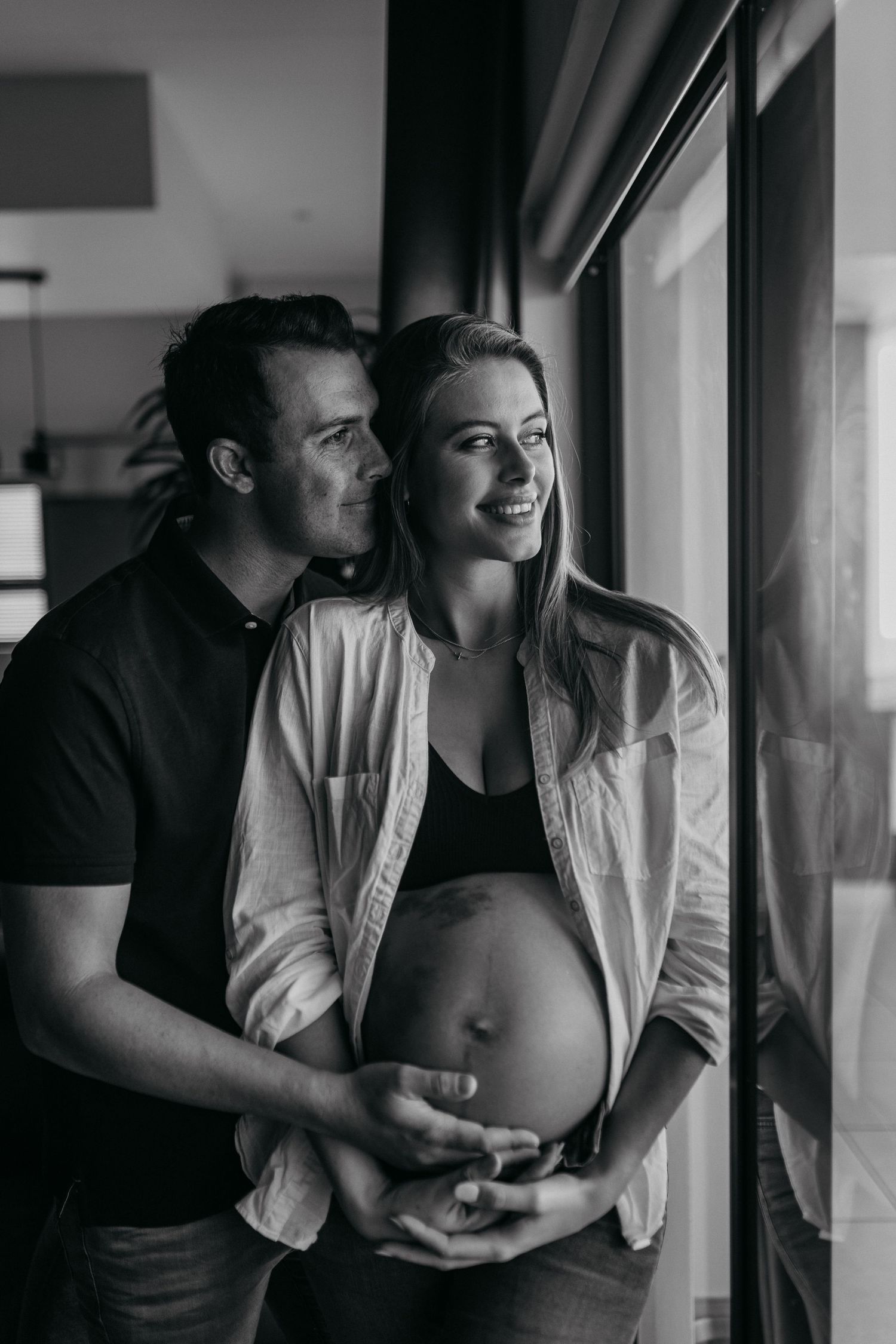 Couple embraces near window in intimate black and white maternity photography session.