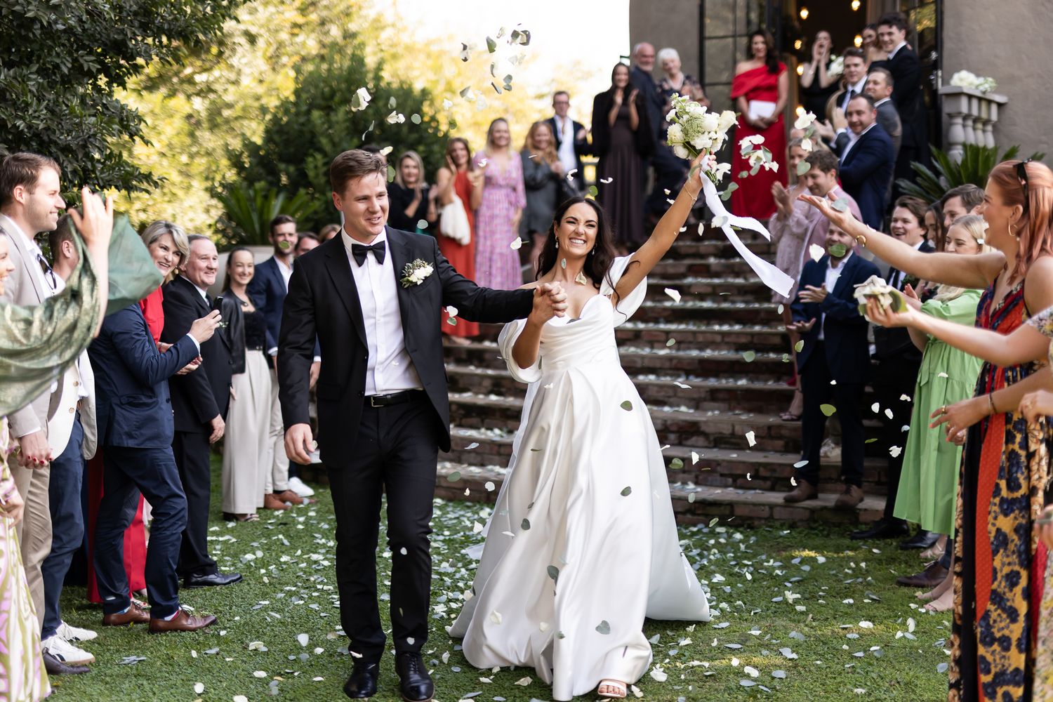 A wedding couple exits their ceremony while guests throw flower petals on a stone staircase outdoors.