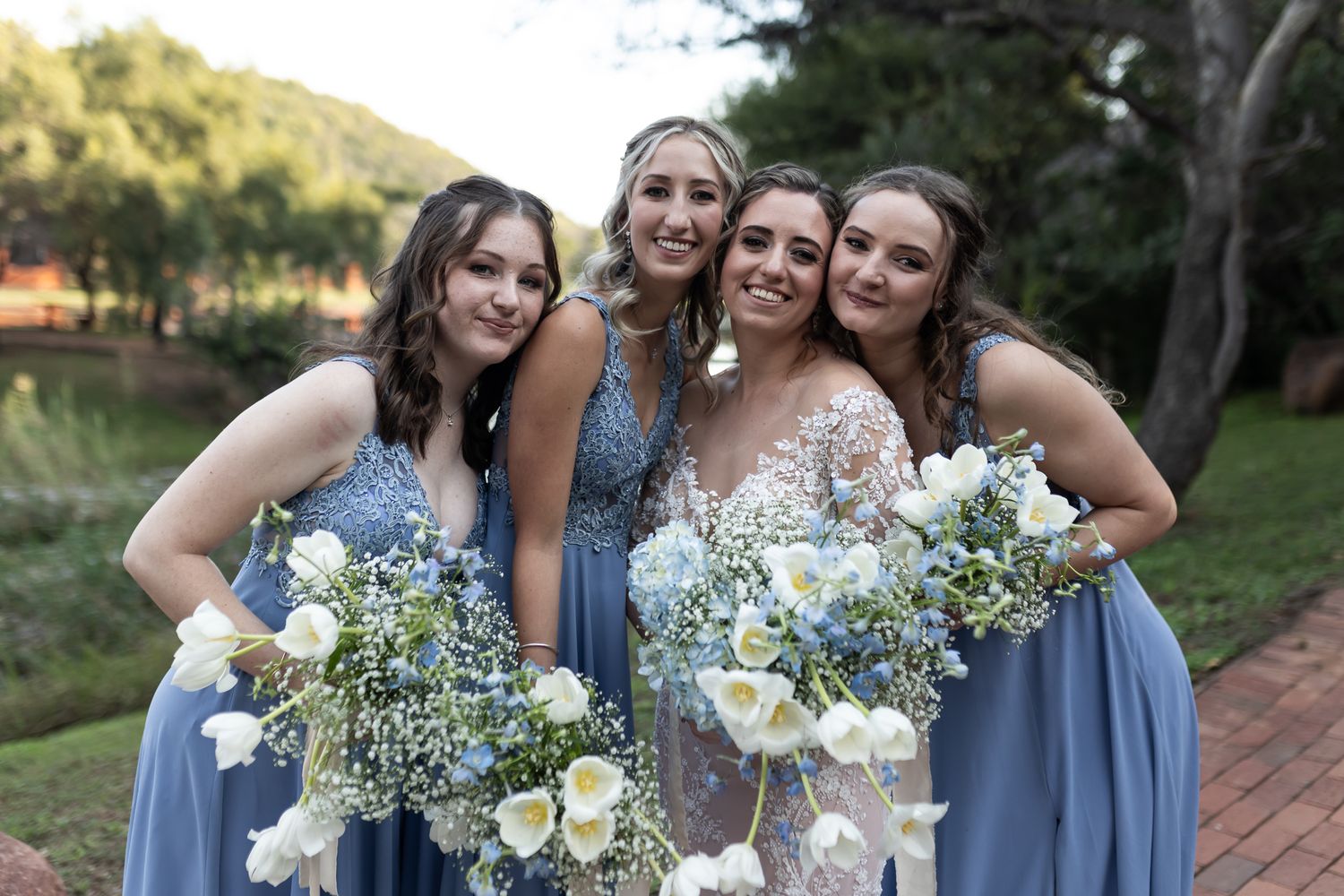Bridesmaids in dusty blue dresses hold white floral bouquets while celebrating outdoors.