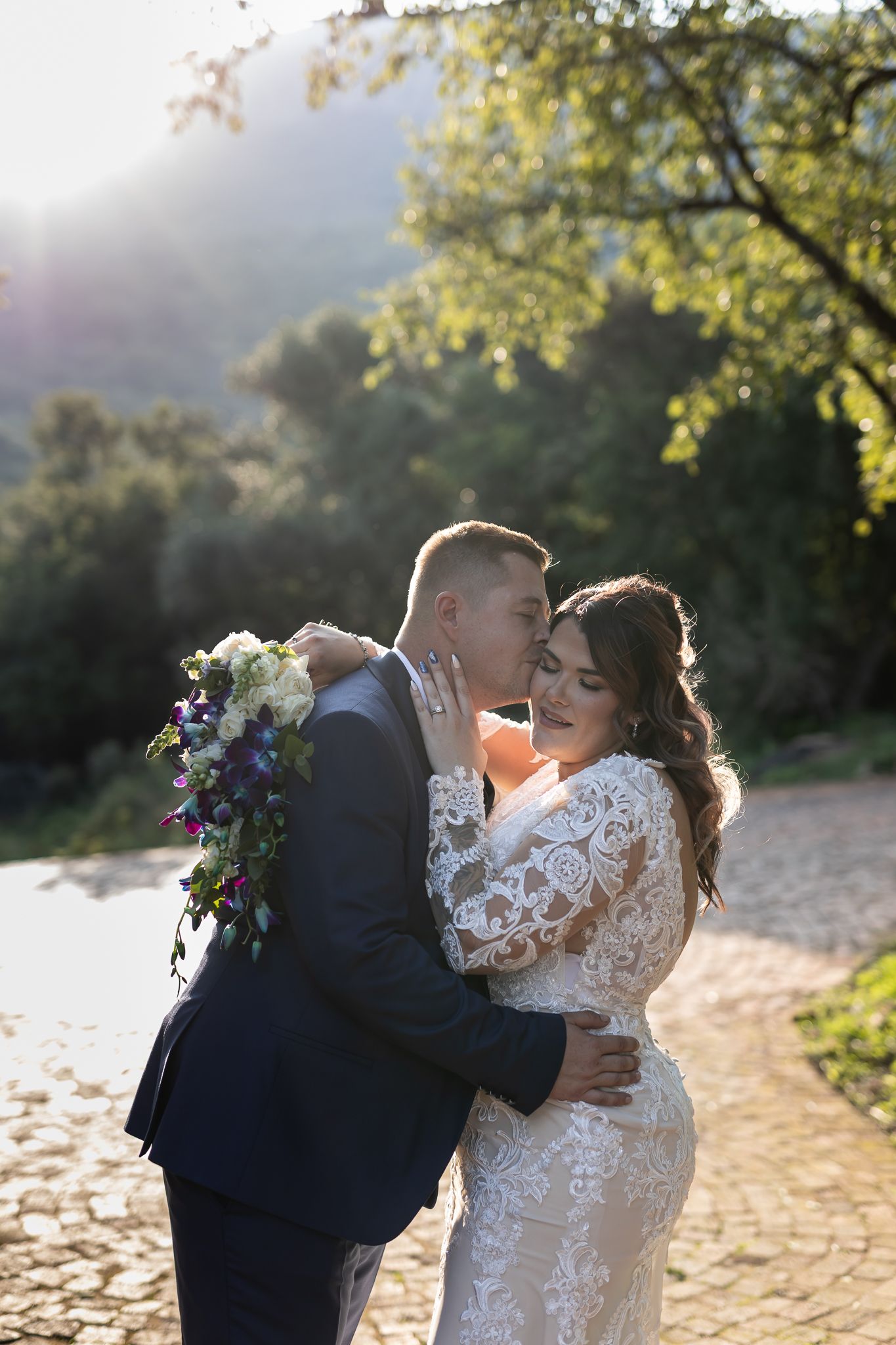Bride in lace gown and groom in navy suit embrace during golden hour wedding photos.