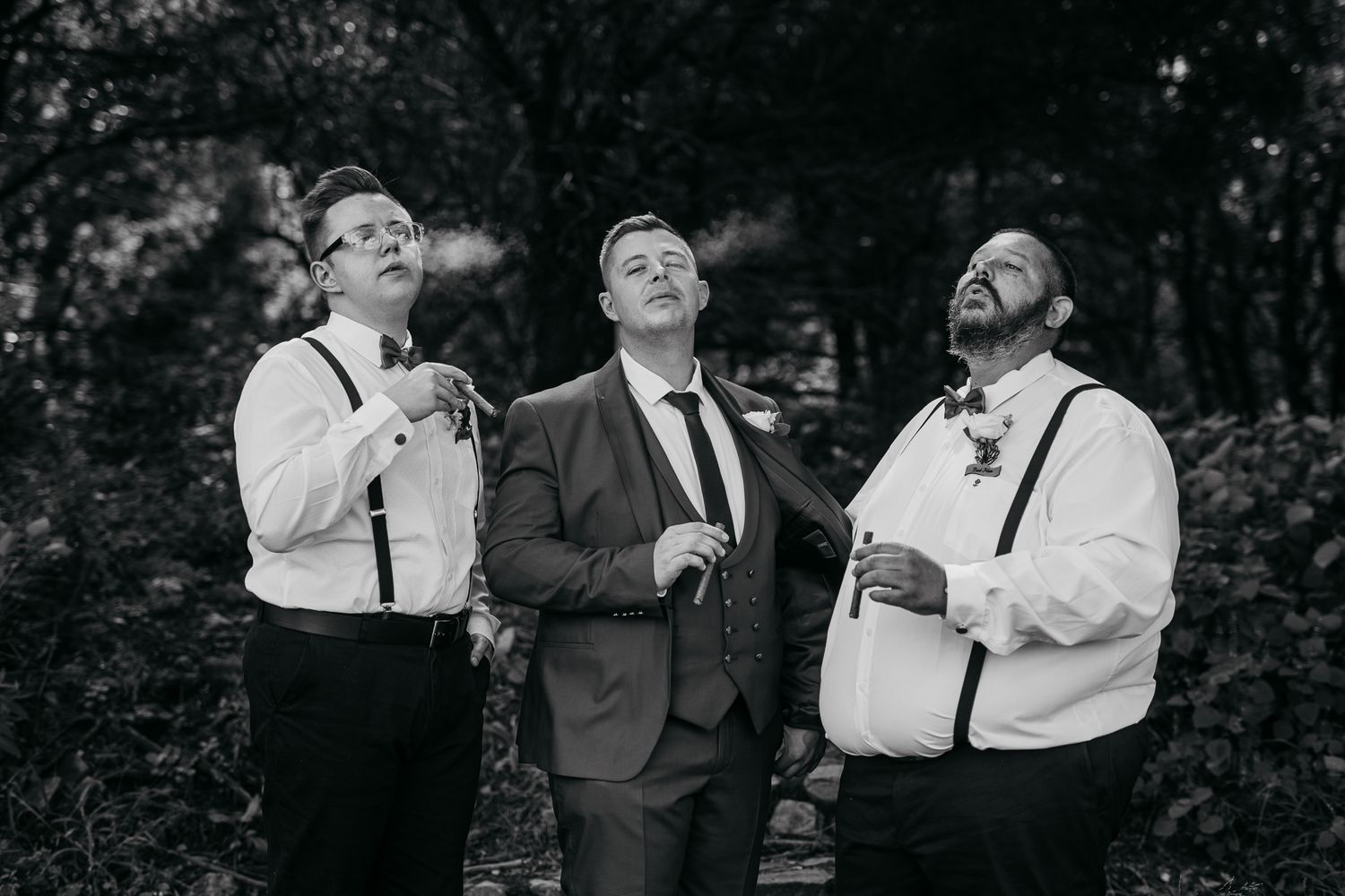 A black and white photo of three groomsmen sharing a candid laugh in a garden setting.