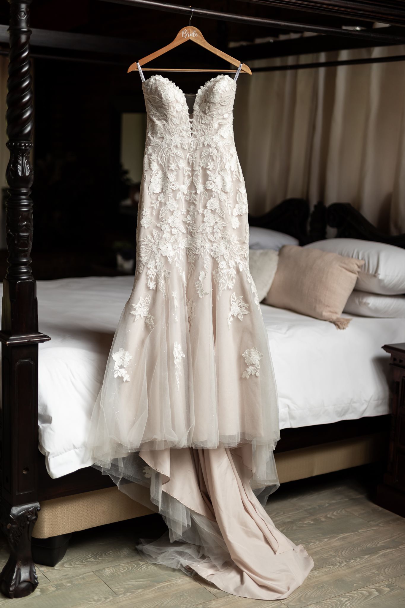 Elegant lace wedding dress hanging on wooden hanger against dark bedroom background.