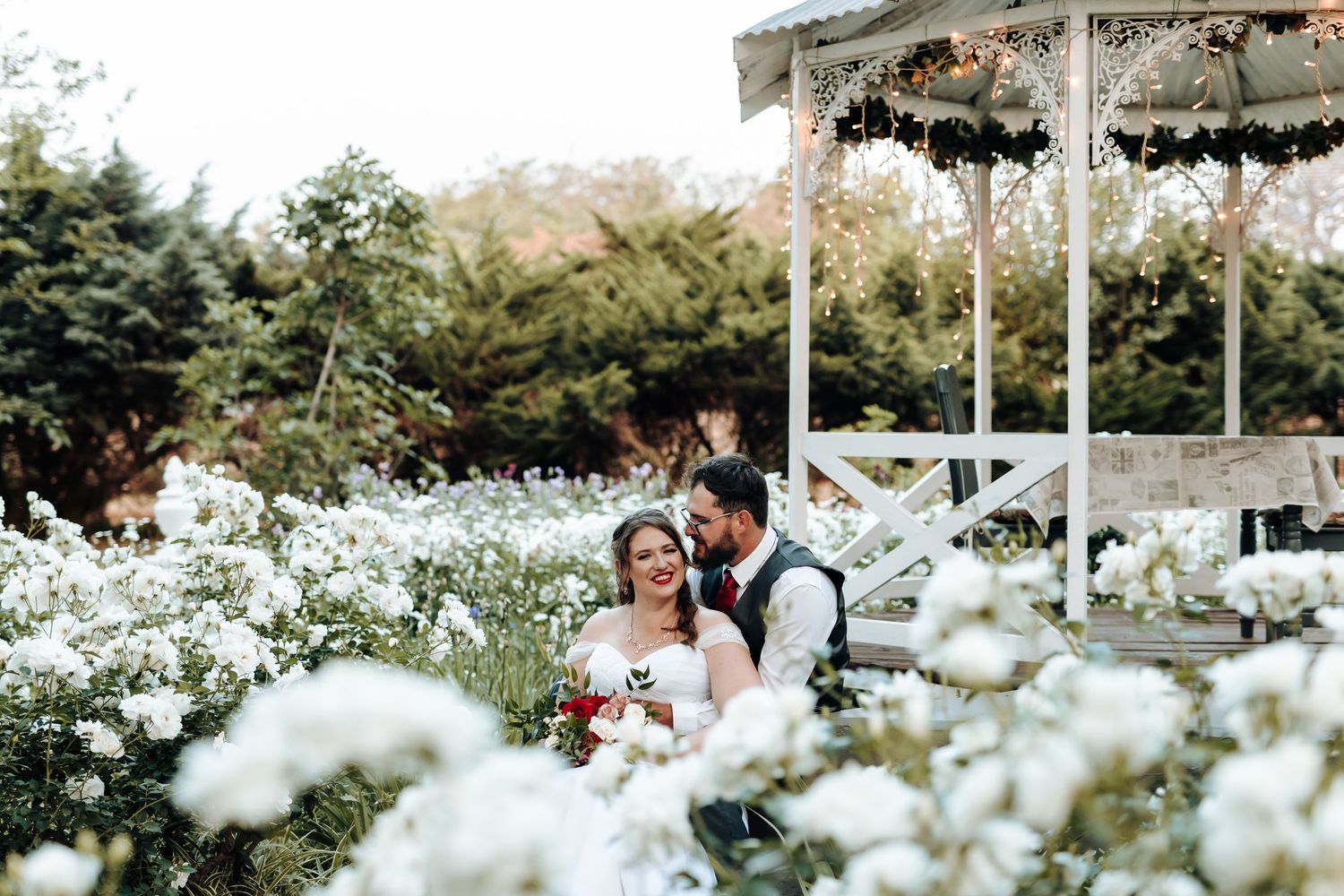 Wedding couple surrounded by white flowers in front of a decorated gazebo.