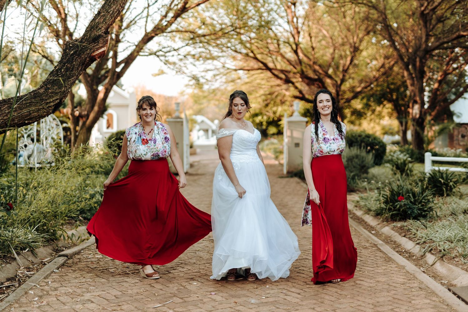 Bridal party in flowing red dresses and white gown pose on brick walkway under autumn trees.