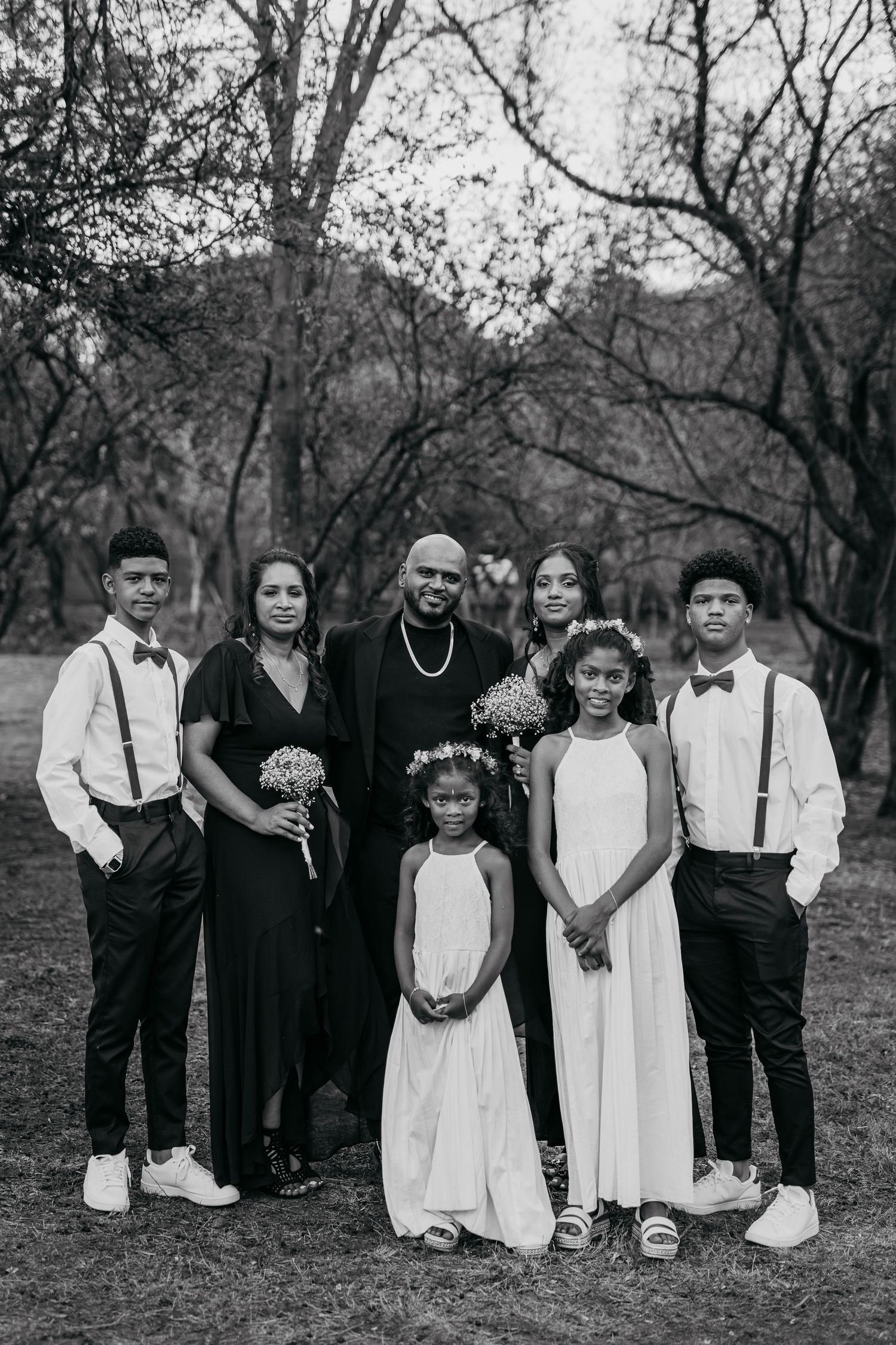 A black and white portrait of a wedding party in formal attire standing together outdoors.