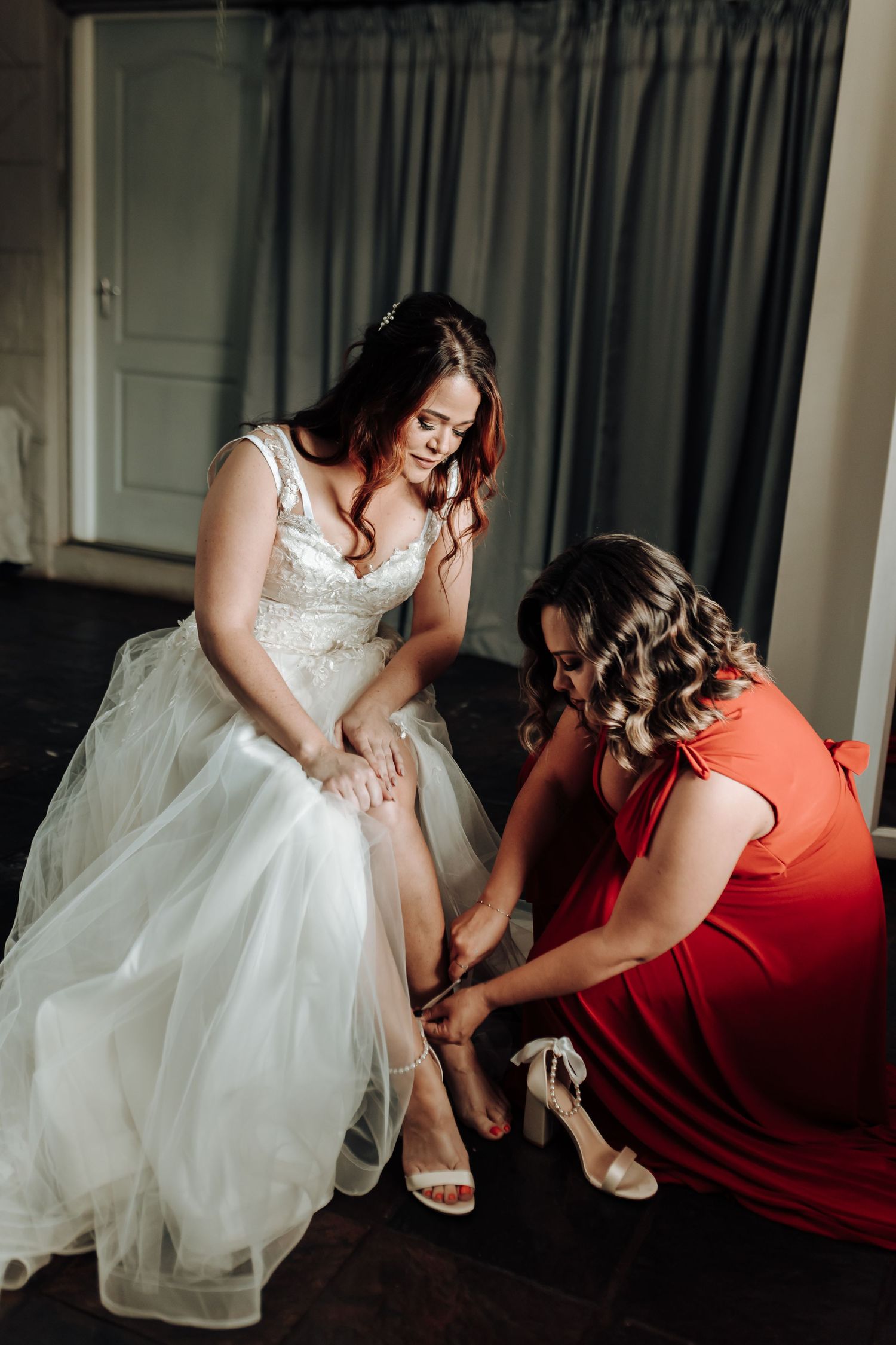 Bridesmaid in red dress helps bride put on elegant white wedding sandals.