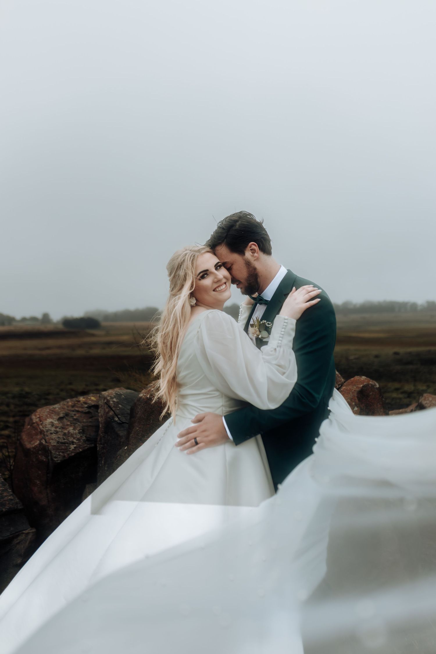 Intimate wedding portrait showing a tender moment between newlyweds with flowing white dress against moody landscape.