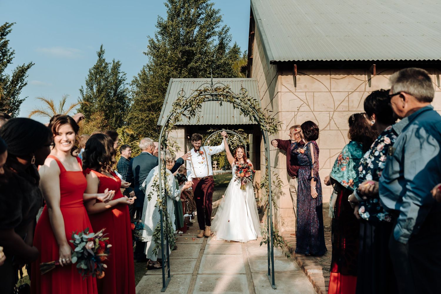 Outdoor wedding ceremony with guests in red dresses watching bride walk down aisle under decorative arch.