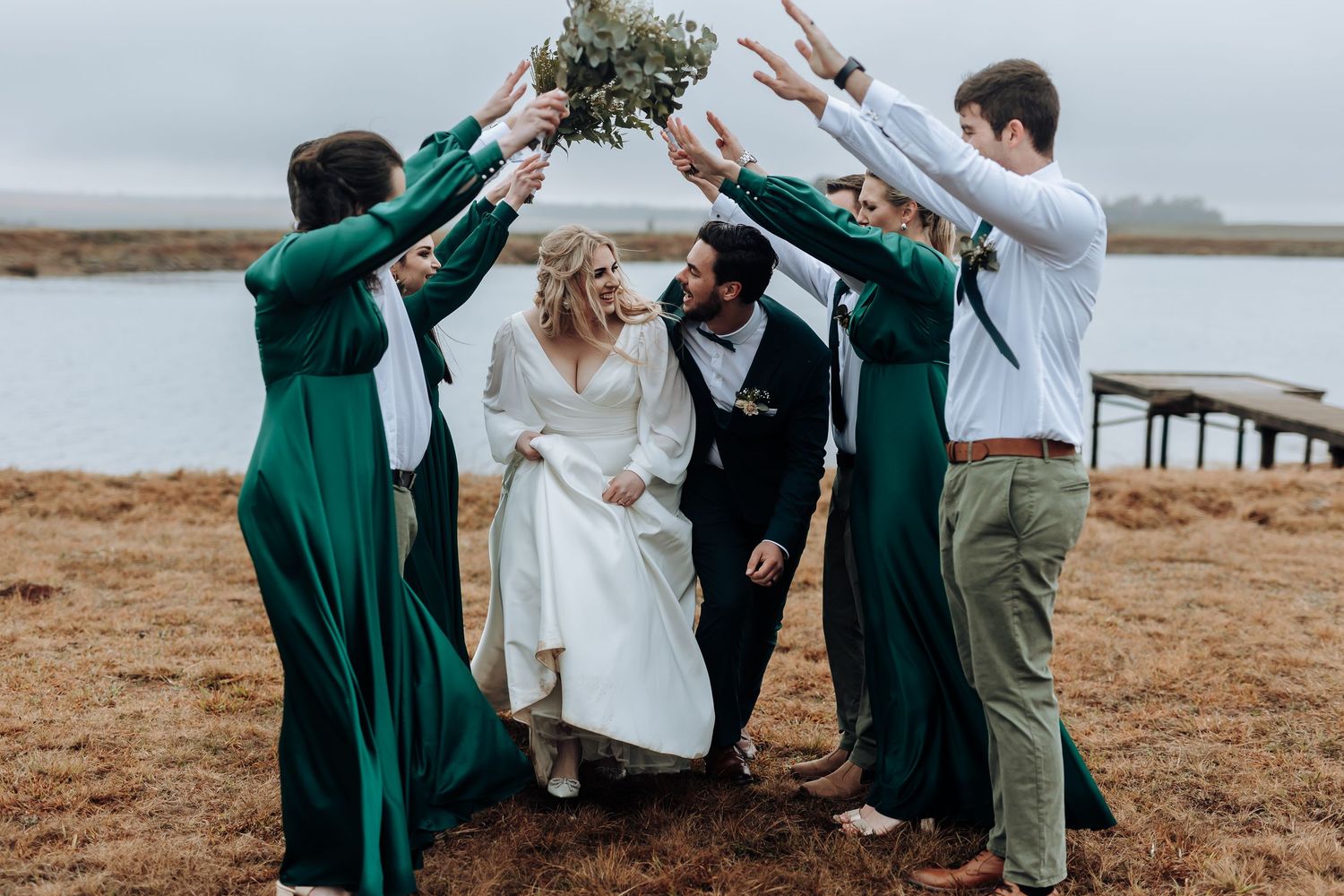 Wedding party forms celebratory arch with bouquet over couple by waterfront.