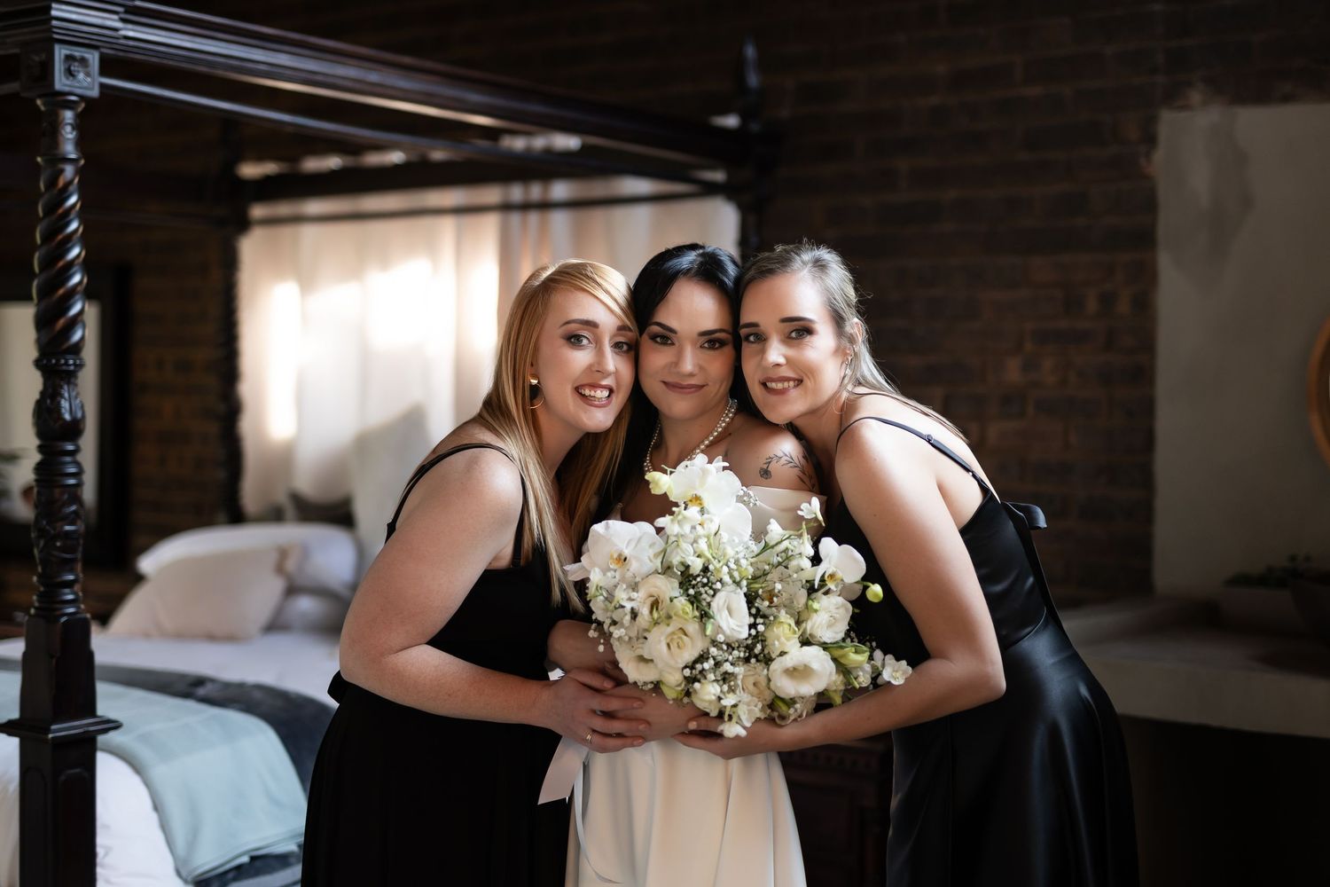 Three women in formal dresses pose together holding a white flower bouquet in an indoor setting.