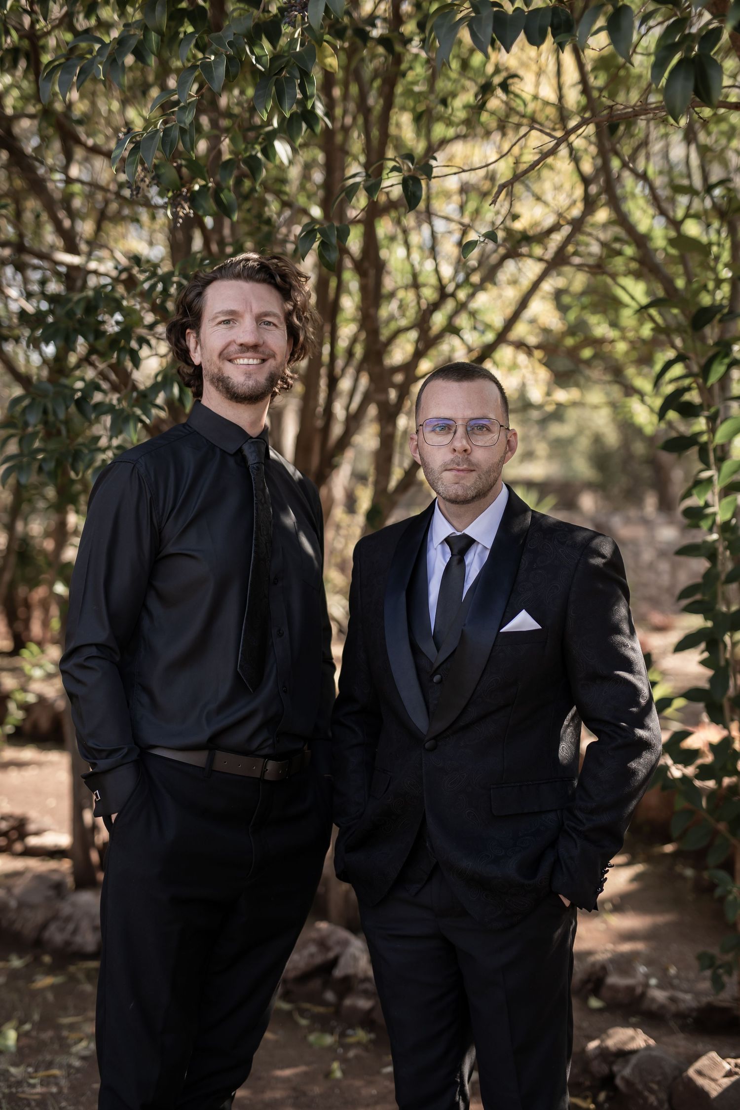 Two men in formal black suits posing together in an outdoor garden setting.