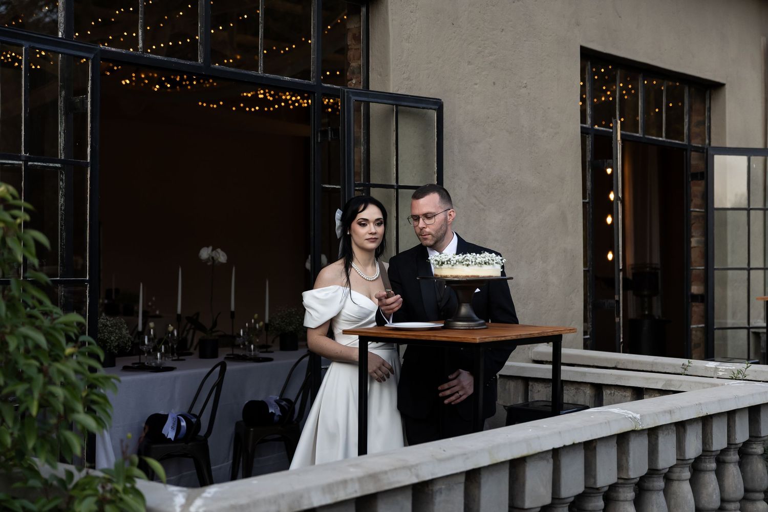 A couple in wedding attire standing on a balcony with string lights while looking at a menu together.