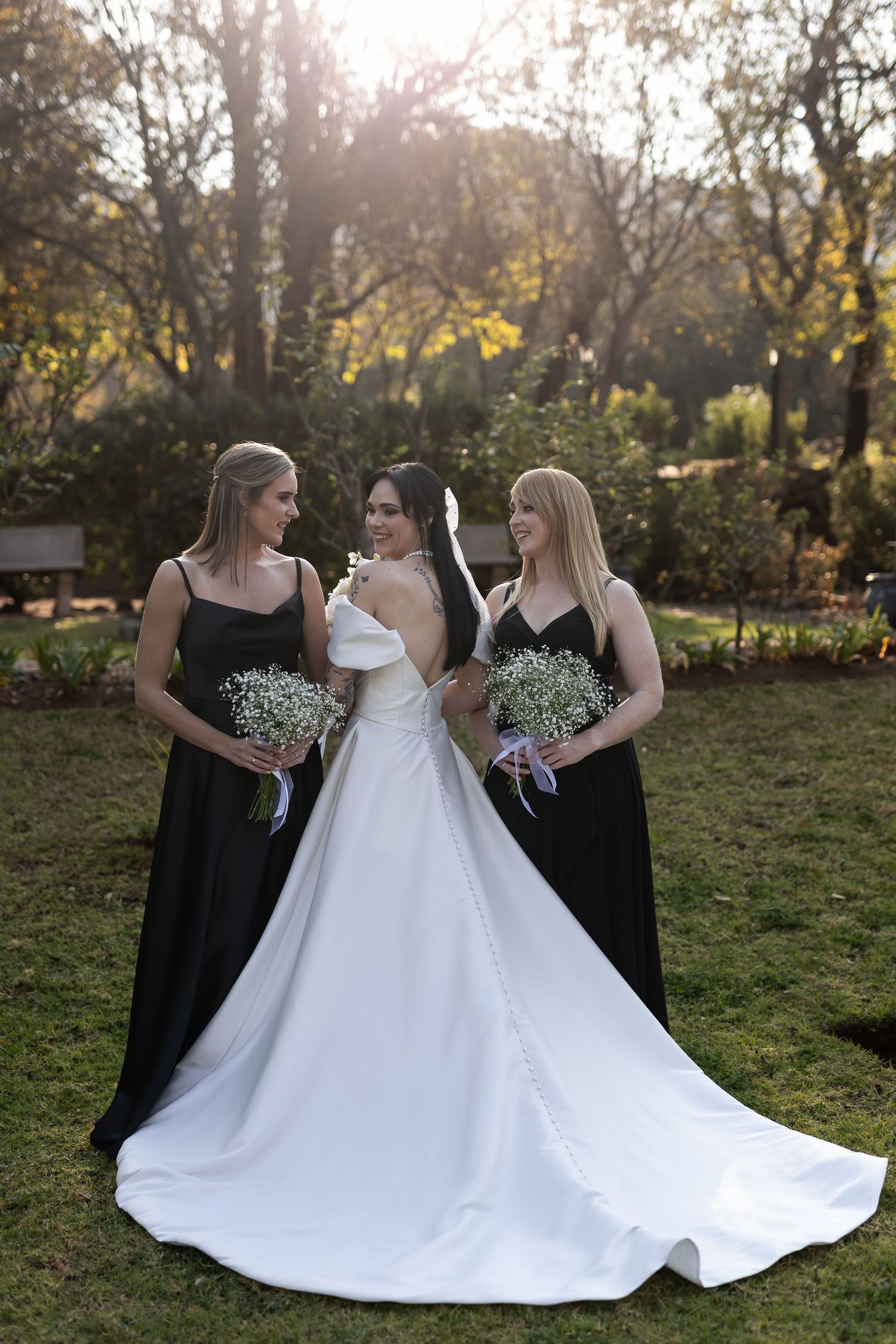 Three women in a garden setting wearing black bridesmaid dresses and white wedding gown for outdoor wedding ceremony.