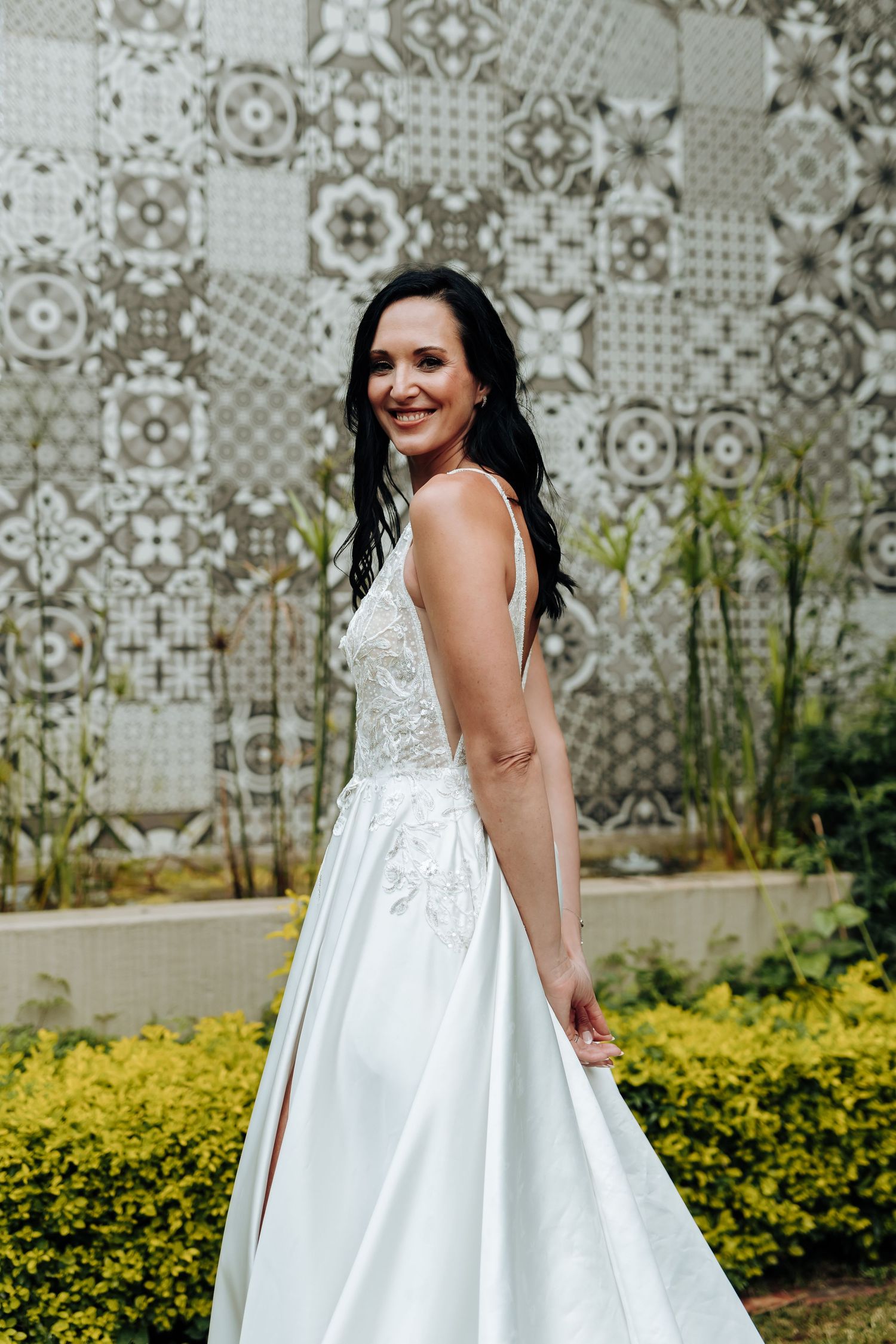 Smiling bride in a white lace A-line wedding gown posing outdoors against a decorative tiled wall.