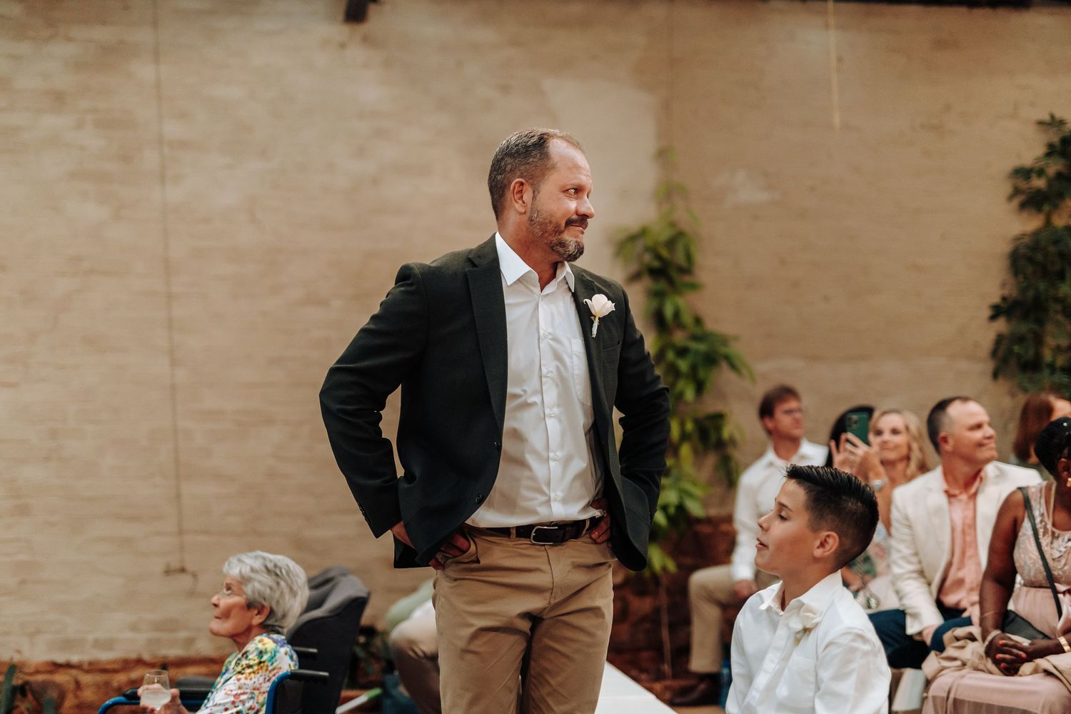 Groom in navy blazer and khaki pants smiling at a young boy during an outdoor wedding ceremony.