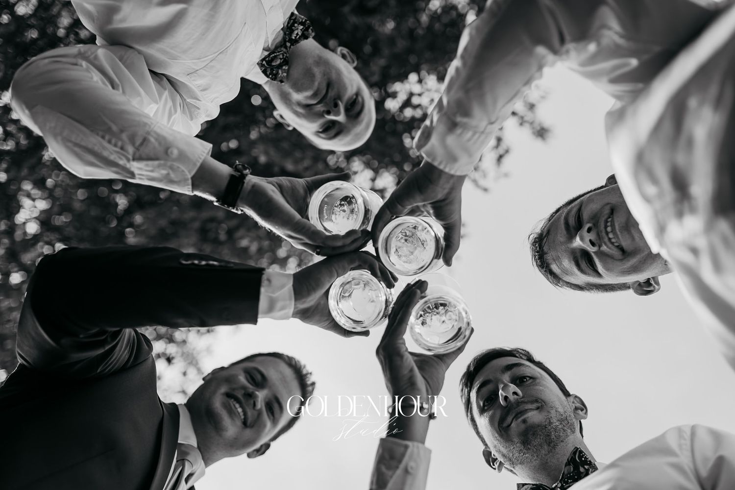 Fun low-angle black and white shot of groomsmen toasting with glasses at an outdoor wedding celebration.