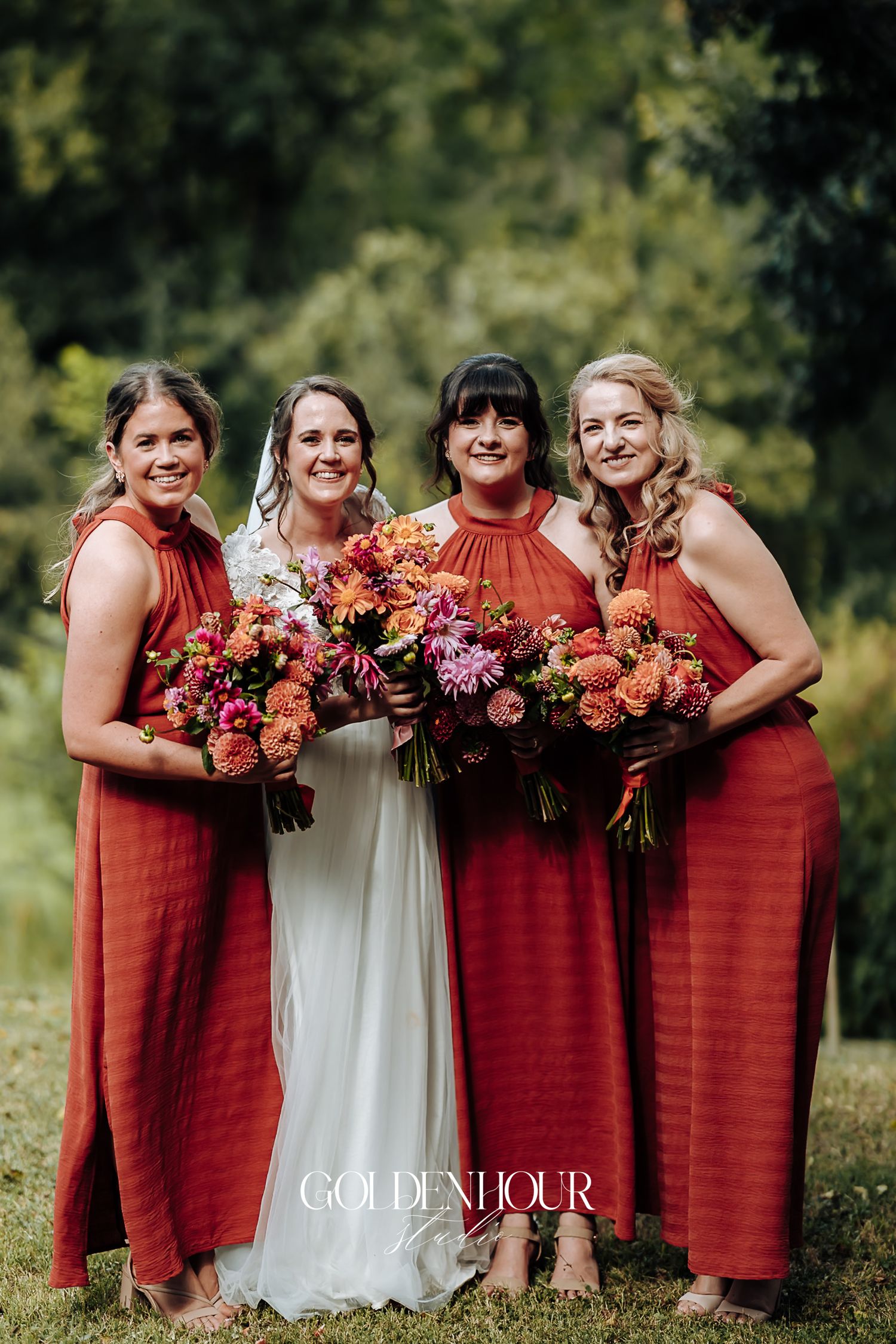 Bride posing with three bridesmaids in terracotta dresses holding colorful bouquets outdoors.