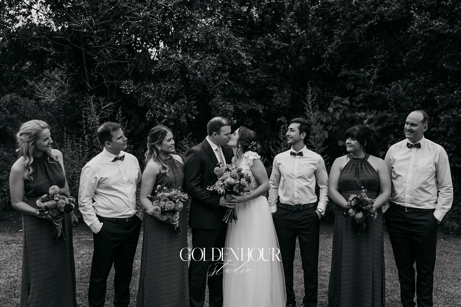 Black and white wedding party photo with bride and groom kissing flanked by bridesmaids and groomsmen.