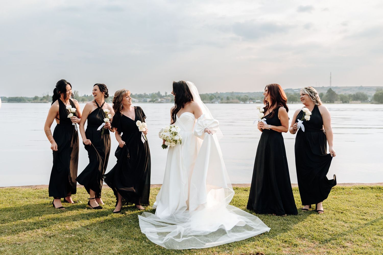 A bridal party in black dresses and white gown sit together on grass overlooking a scenic waterfront view.