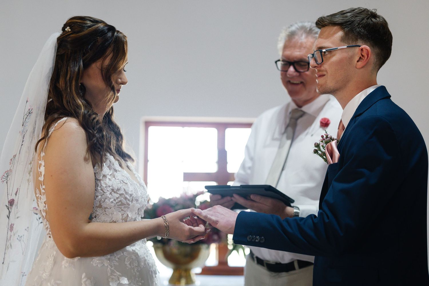 Wedding couple exchanges rings during intimate indoor ceremony with natural lighting from nearby window.