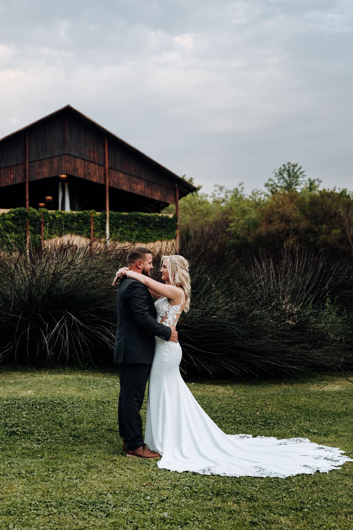 A romantic wedding couple dances together on a green lawn with a rustic barn in the background.