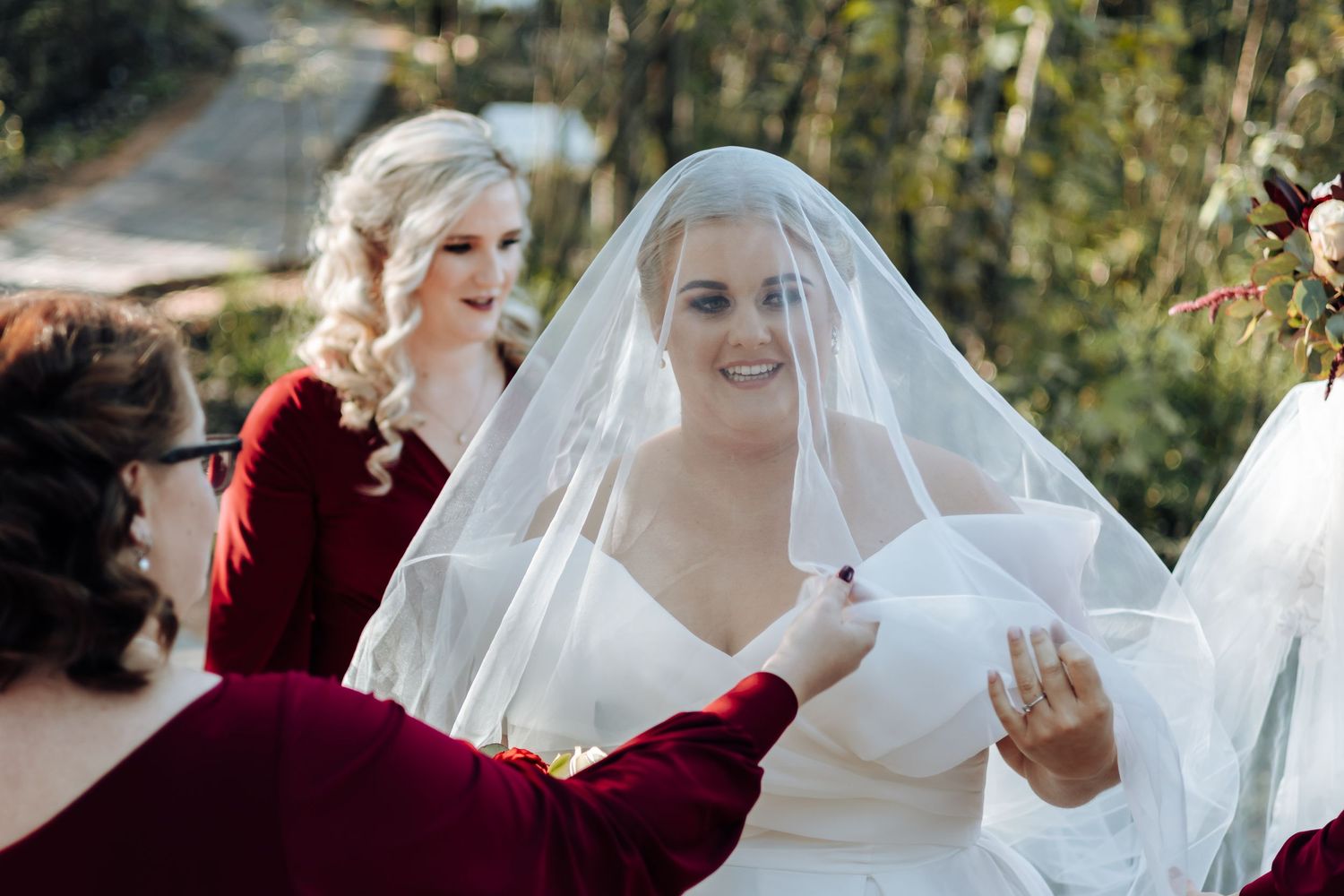 Bride smiling under veil while bridesmaid in burgundy dress adjusts it outdoors at wedding ceremony.