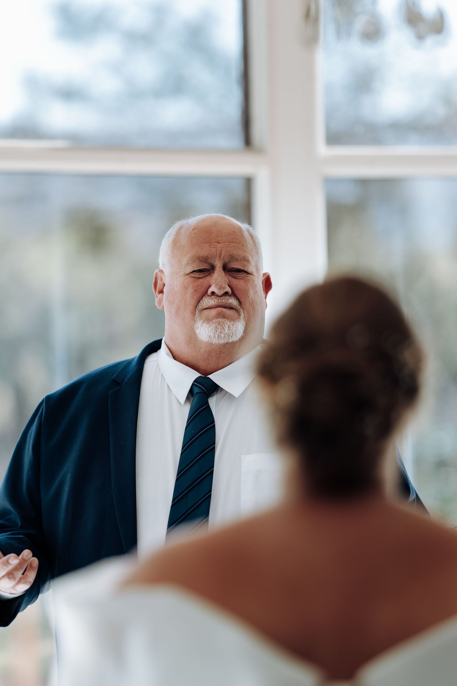 Emotional father sees bride daughter for first time on wedding day, standing near large window in suit and tie.