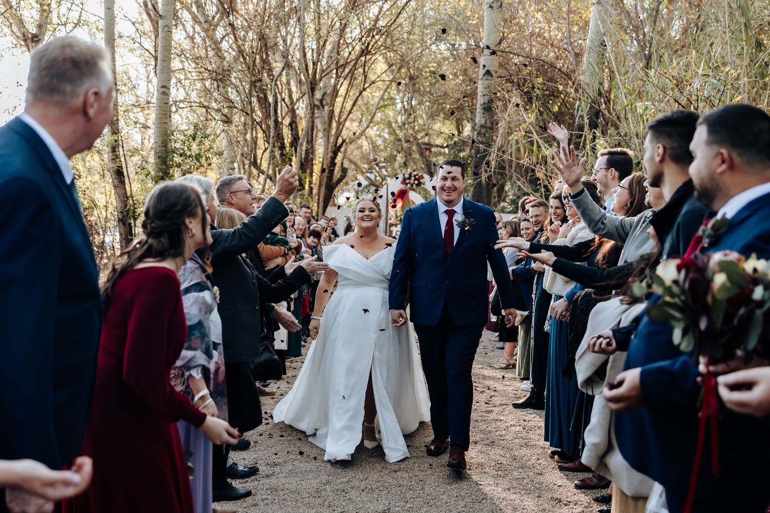 Joyful newlyweds walk hand-in-hand through a celebratory outdoor aisle lined with cheering guests at a woodland wedding.