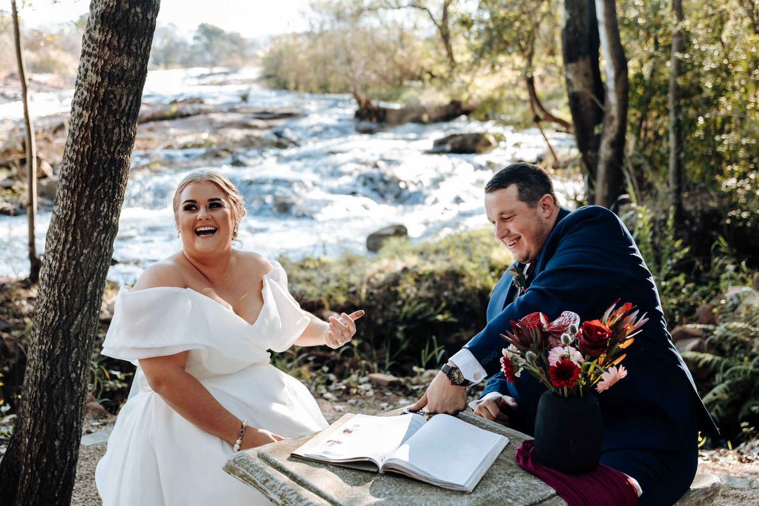 Joyful bride and groom signing wedding registry outdoors by a scenic waterfall, bride in white off-shoulder gown.
