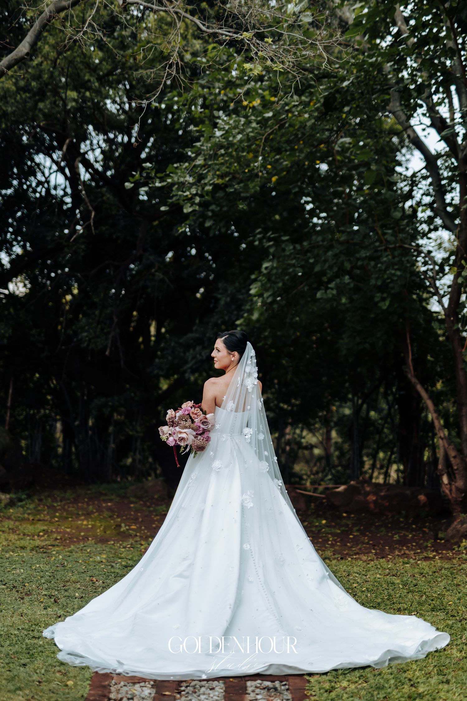 Stunning bride in a flowing white gown with floral veil holding a pink bouquet in a lush garden setting.