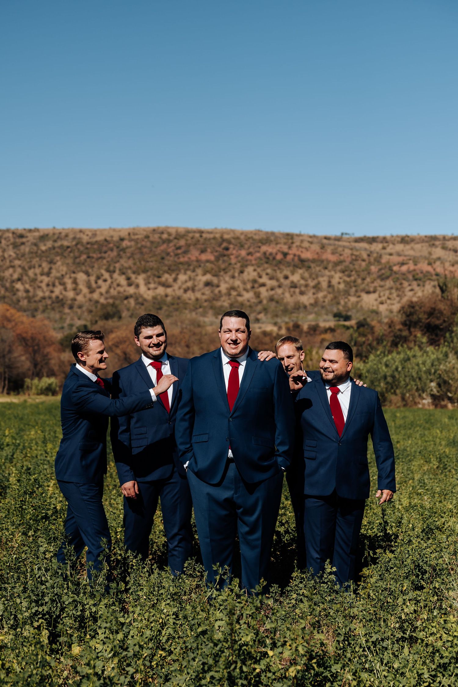 Groom and groomsmen in navy suits walk through green field with rocky hills under clear blue sky.