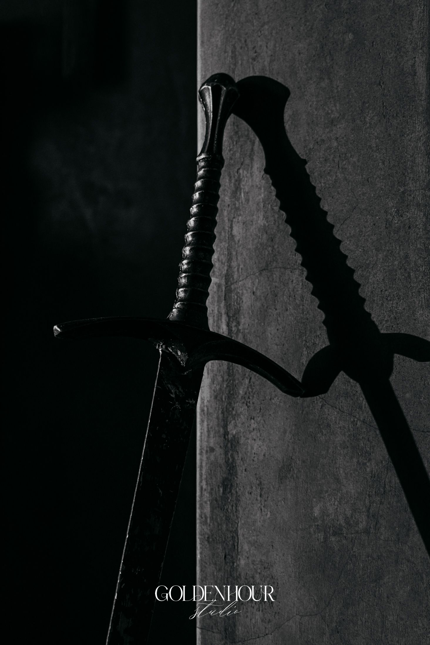 Dramatic black and white close-up of ornate medieval sword leaning against stone wall casting shadow.