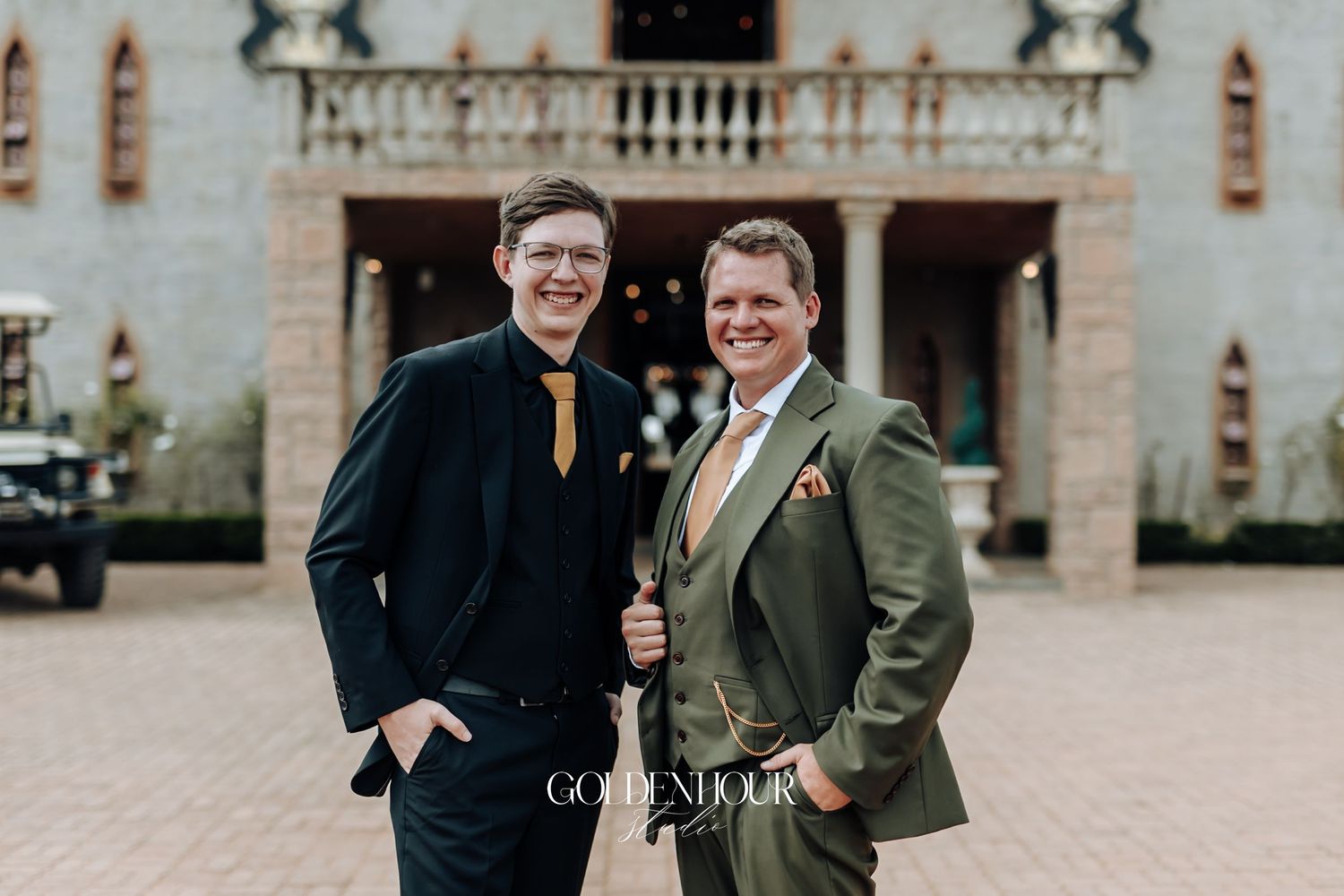 Two grooms in navy and olive suits smiling outside ornate wedding venue facade.