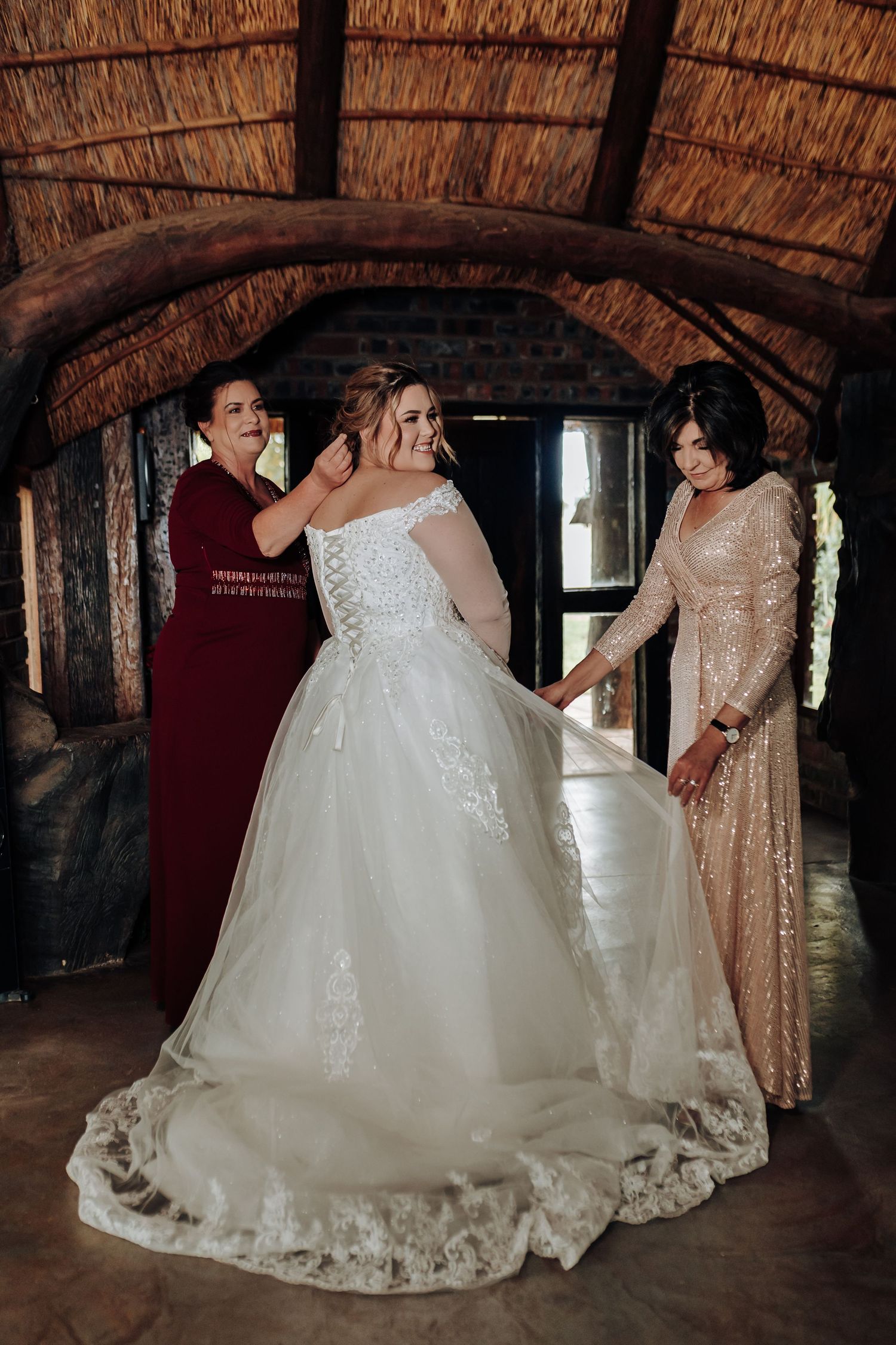 Three people in formal attire gather in a rustic thatched-roof venue with one wearing an elegant white wedding gown.