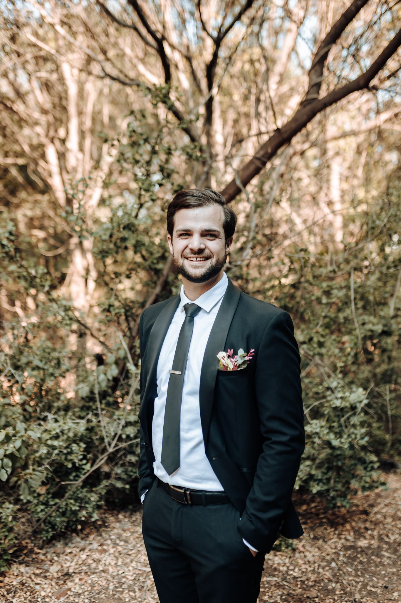 Sharp suited figure stands casually in a wooded area with sunlit trees in the background during a formal event.