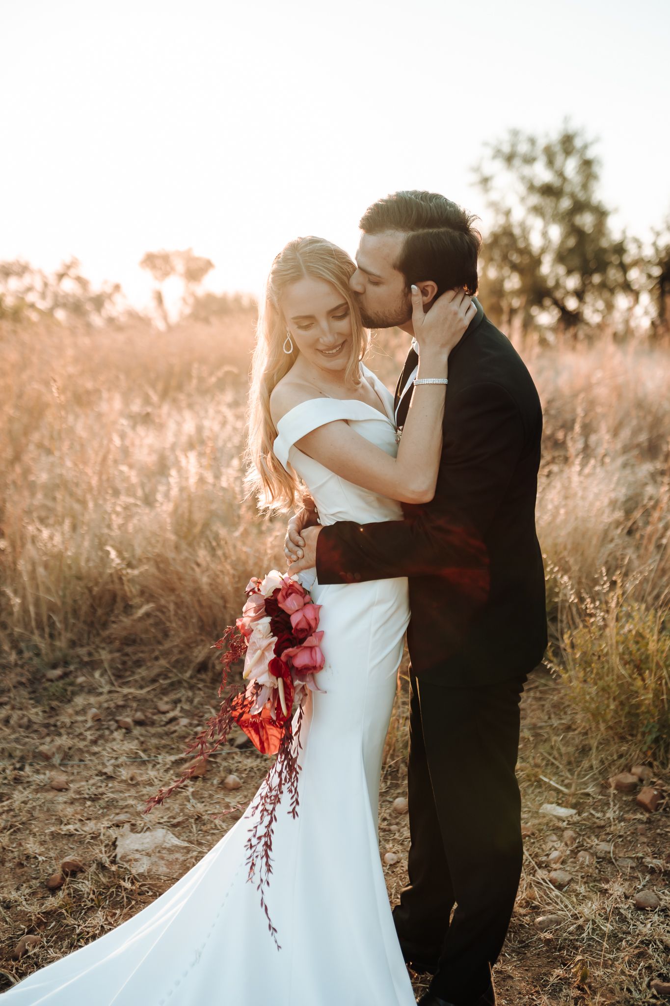 A romantic sunset wedding portrait of a couple embracing in a golden field with the bride holding a cascading bouquet.