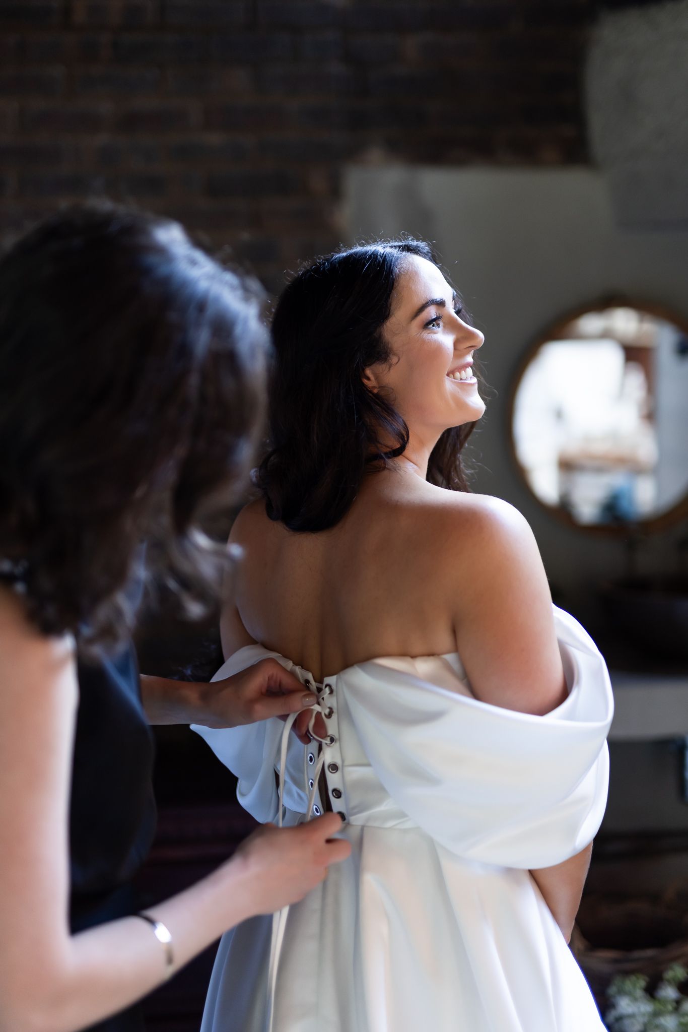 Wedding dress being laced up in brick-walled getting ready room.