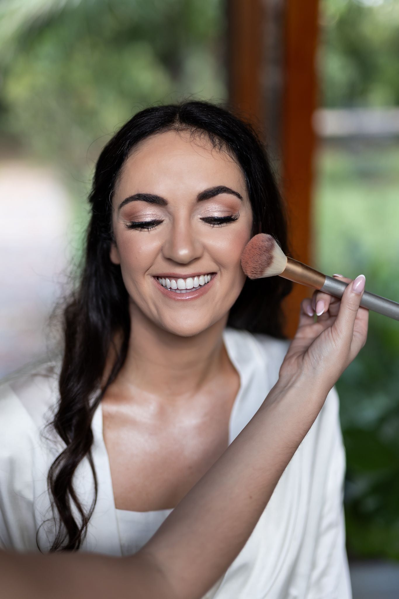 Woman having makeup applied with brush while smiling during wedding preparation.