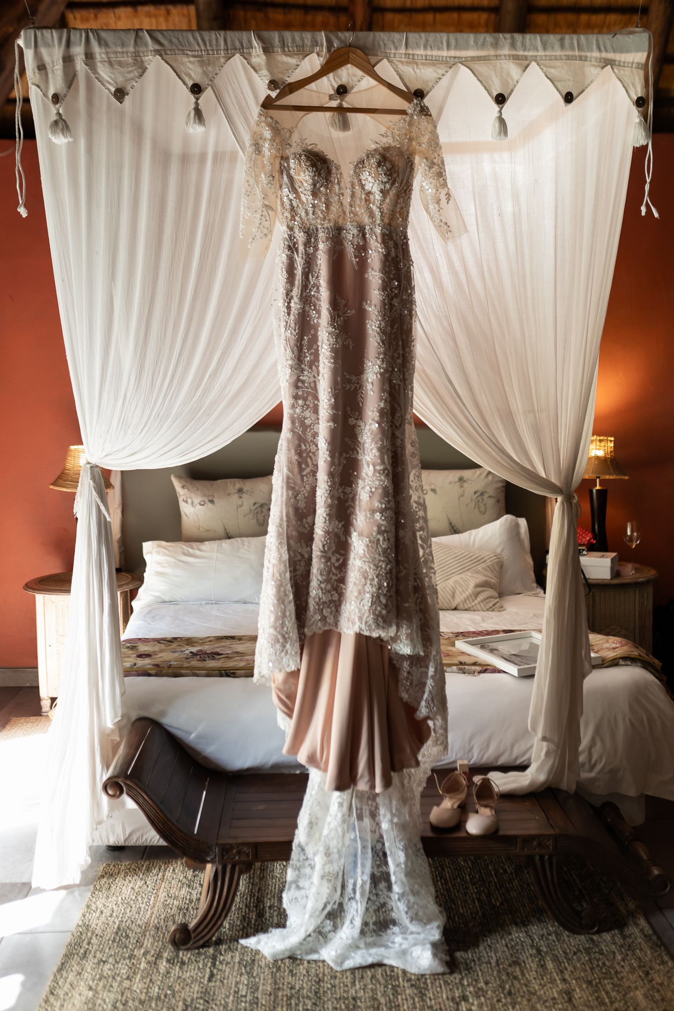Elegant lace wedding dress hanging from canopy bed with white curtains.